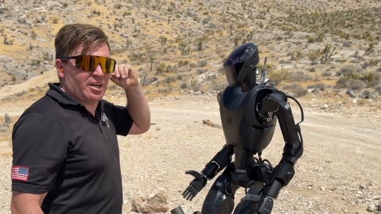A screenshot from a Foundation video shows one of the company's humanoid robots in a desert environment, alongside a man wearing a black polo and sunglasses.