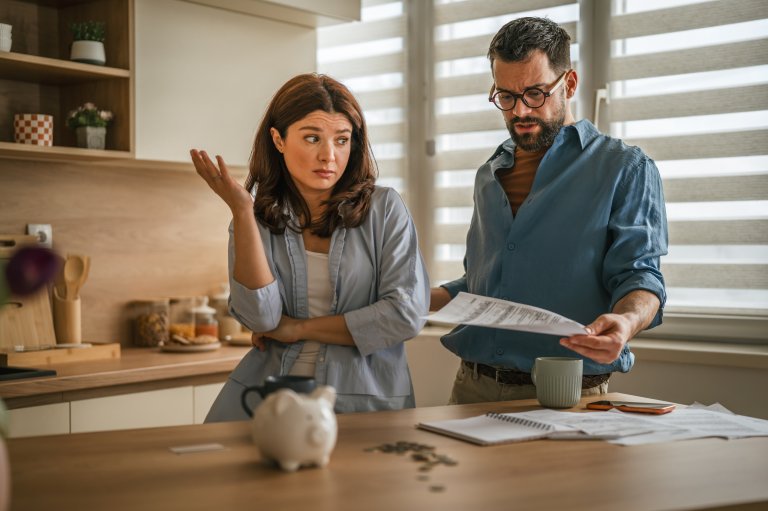 A photo illustration shows a couple being stressed out about bills, with a piggy bank surrounded by a meager amount of coins.