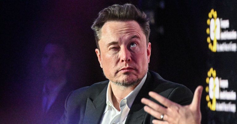 A man with short, dark hair and a light beard is wearing a white shirt and a dark blazer. He is winking with his left eye and looking slightly upward. A blurred hand is visible in the foreground, gesturing. The background is dark with some yellow and white text partially visible on the right side.
