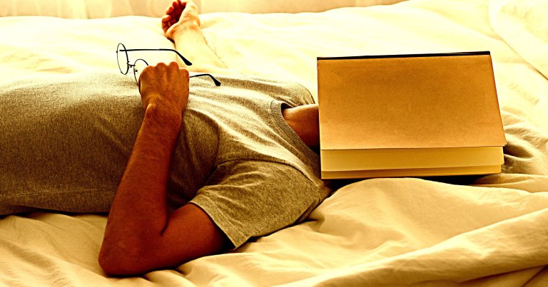 A photo of a man lying on a bed with a book on his face.