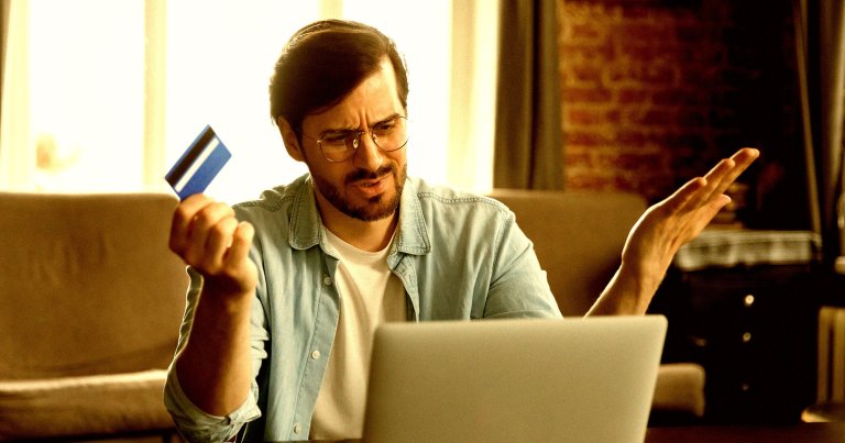 A photo of a man looking shocked at a computer while holding a credit card.