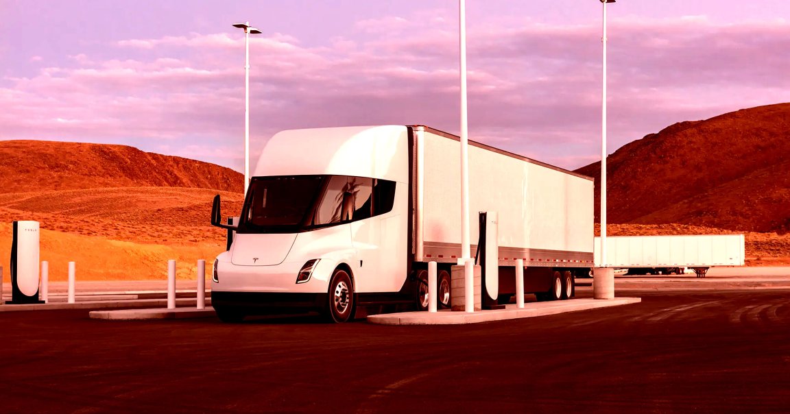 A white Tesla electric semi-truck is parked at a charging station in a desert-like area with reddish-brown hills in the background. The sky is a soft pinkish-purple with scattered clouds. The truck has a sleek, modern design with a large windshield and smooth contours. The charging station features Tesla-branded chargers and tall light poles.