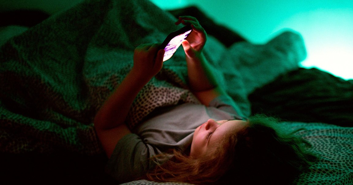 A child lying in bed under a patterned blanket, holding and looking at a glowing smartphone screen in a dimly lit room with a greenish-blue ambient light.