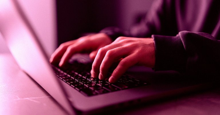 Hands typing on a laptop keyboard with a pinkish-purple lighting effect, wearing a dark long-sleeve garment.