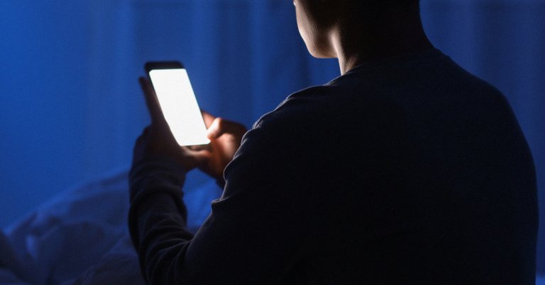 A man texting on a smartphone in a dark bedroom.