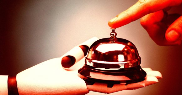 A gloved hand is holding a shiny service bell while a bare finger is pressing down on the top of the bell. The bell is metallic and reflective, and the glove is white with a dark band around the wrist. The background is softly blurred.