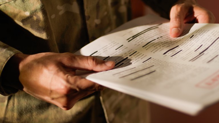 Hands holding a document with multiple lines of text, many of which are blacked out or redacted, suggesting it is a classified or confidential paper. The person holding the document is wearing a camouflage-patterned uniform.