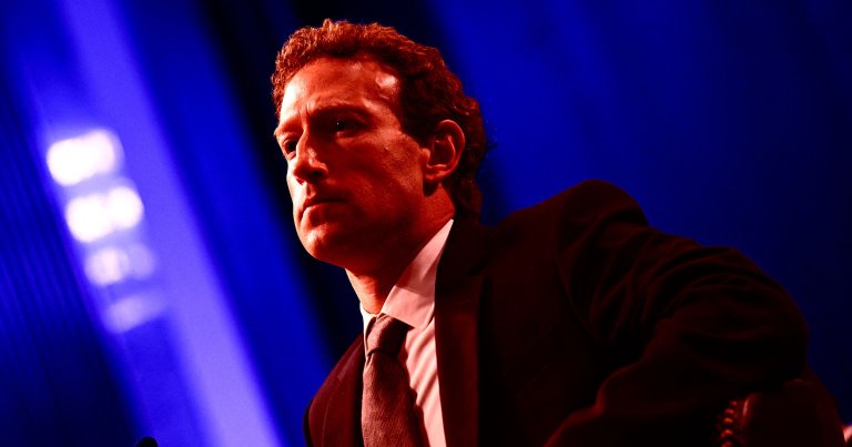 A man with curly hair wearing a dark suit, white shirt, and tie is shown in a close-up shot with dramatic red lighting on his face and upper body. The background is a deep blue with blurred light patterns. The man appears serious and focused.