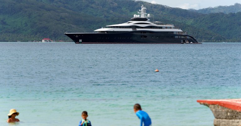 A large, sleek black and white luxury yacht is anchored in calm blue waters near a lush, green mountainous coastline. In the foreground, three people, including two children and an adult wearing a hat, are enjoying the shallow water near the shore. The yacht features multiple decks with large windows and modern design elements.