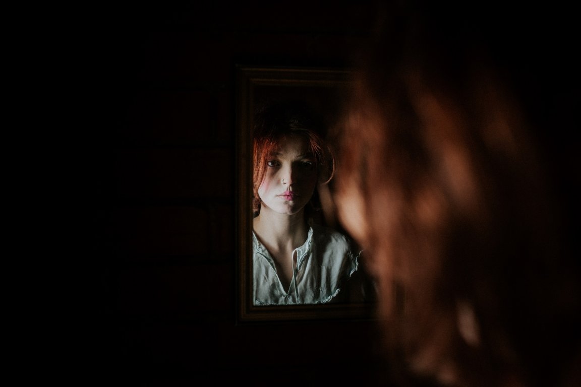 A young woman with red hair and a pale green blouse is looking at her reflection in a small, dimly lit mirror. The background is dark, and her face is partially illuminated, showing a serious expression. The image captures her reflection clearly, while the back of her head is blurred in the foreground.