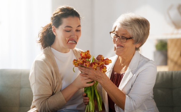 A younger woman and an older woman sitting together, holding a bouquet of orange and yellow tulips. Both have distorted, exaggerated mouths that appear digitally altered, creating an unusual and humorous effect. The younger woman has dark hair tied back and is wearing a beige cardigan over a white shirt, while the older woman has short gray hair, glasses, and is dressed in a white textured jacket over a maroon top. They are indoors with soft natural light.