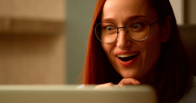 A close-up of a woman with long red hair and round glasses, looking at a screen with an expression of surprise and delight. Her mouth is open in a smile, and her hand is resting near her chin. The background is softly blurred.