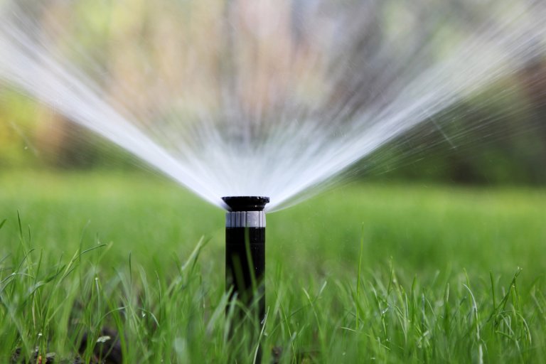 A black garden sprinkler is spraying water in a wide arc over green grass. The water streams are captured in motion, creating a fan-like pattern. The background is blurred with natural colors, emphasizing the sprinkler and the water spray.