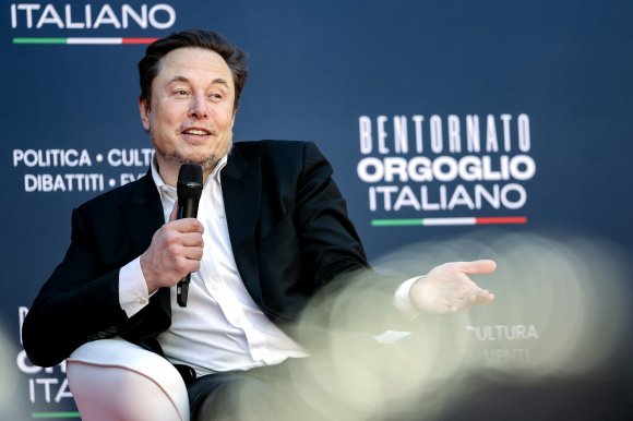 Elon Musk is seated in a white chair, holding a microphone in his right hand and gesturing with his left hand. He is wearing a black suit jacket over a white shirt. The background features Italian text and the colors of the Italian flag.