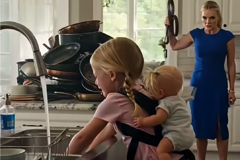 A young girl with braided blonde hair is washing dishes at a kitchen sink, carrying a baby on her back in a baby carrier. The baby has a small ponytail and is wearing a light-colored outfit. In the background, a woman in a blue dress stands with an intense expression, holding a belt raised in one hand. The kitchen counter behind the girl is cluttered with a large stack of dirty pots and pans.