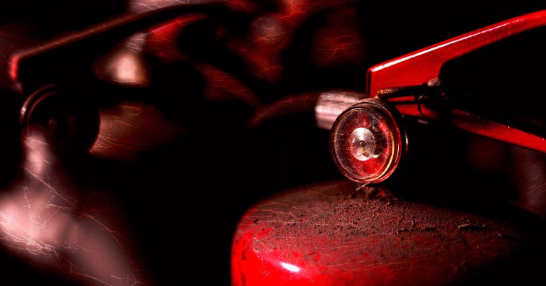 Close-up of a red fire extinguisher valve and pressure gauge, with a dark, moody background and visible scratches on the surface.