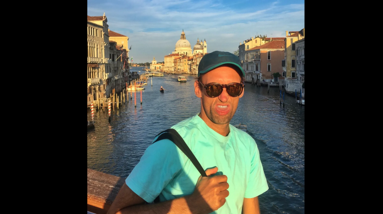A man wearing a light turquoise t-shirt, dark sunglasses, and a dark green cap stands on a bridge over a canal. He is holding a black strap over his shoulder and making a grimacing facial expression. The background shows a canal with boats and gondolas, flanked by historic buildings with warm sunlight, and a large domed church in the distance under a partly cloudy blue sky.