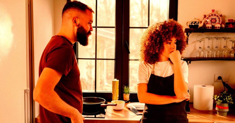 Young pensive woman with curly hair wearing apron looking away and ignoring her boyfriend.