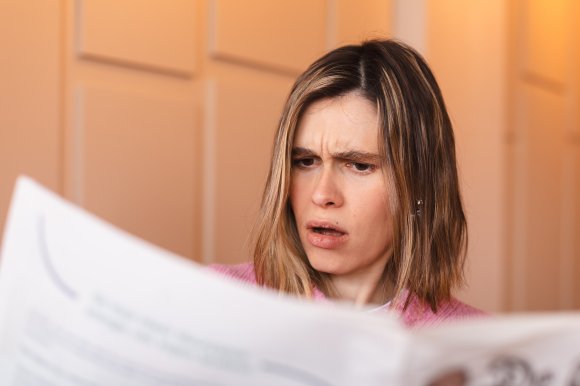 A woman with shoulder-length brown hair and light highlights is wearing a pink sweater and looking at a newspaper with a confused or concerned expression. The background is softly lit with warm tones and features a paneled wall.