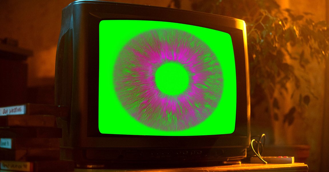 A vintage CRT television displaying a large, stylized eye with a bright green background and a purple iris. The TV is placed on a wooden surface with a warm, dimly lit background featuring some plants and stacked VHS tapes.