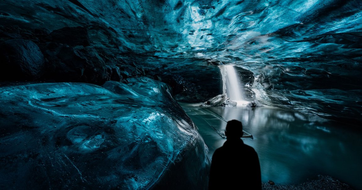 A silhouette of a person stands inside an ice cave with smooth, blue-toned ice walls and ceiling. In the background, a bright waterfall flows through an opening in the ice, creating a small pool of water. The scene has a mystical, serene atmosphere with contrasting dark and light areas.