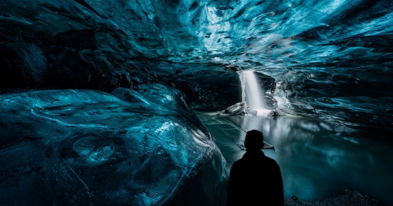 A silhouette of a person stands inside an ice cave with smooth, blue-toned ice walls and ceiling. In the background, a bright waterfall flows through an opening in the ice, creating a small pool of water. The scene has a mystical, serene atmosphere with contrasting dark and light areas.