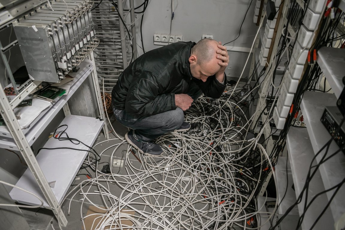 A man is shown crouching on the floor, surrounded by a tangled cables in a room filled with electronic equipment.