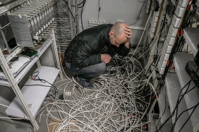 A man is shown crouching on the floor, surrounded by a tangled cables in a room filled with electronic equipment.