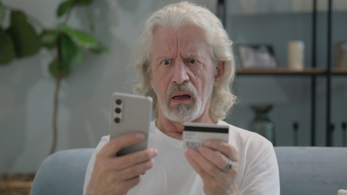 A photo shows an older man looking at a credit card and his phone in confusion.