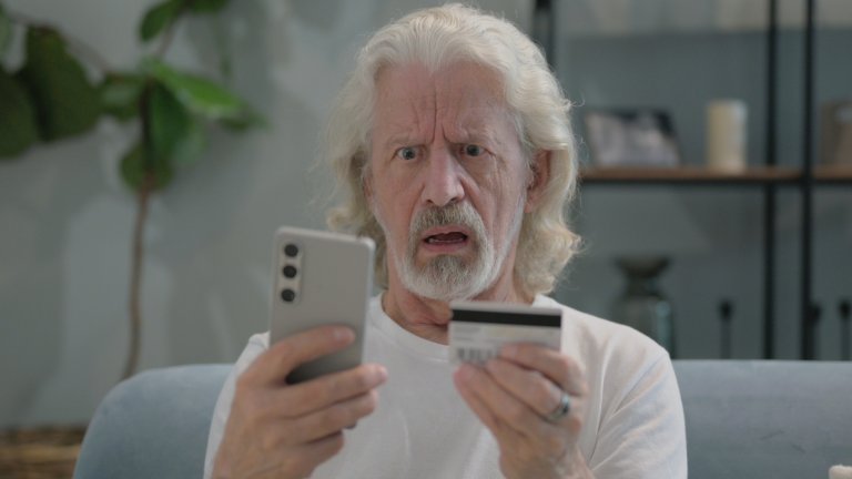 A photo shows an older man looking at a credit card and his phone in confusion.