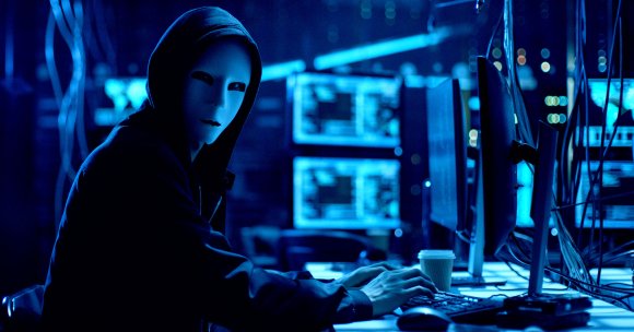A hacker in front of an elaborate computer set up wearing a mask.
