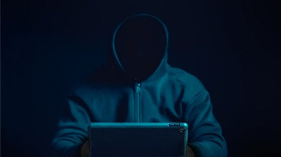 A hooded figure with a hidden face is shown using a computer.