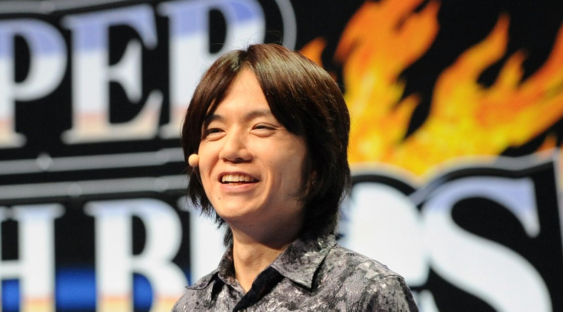 A photo shows Masahiro Sakurai in front of a banner for Super Smash Brothers.